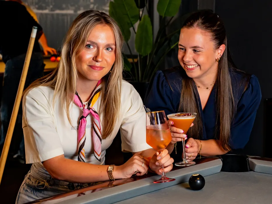 Two women sit at a pool table with a drink, while one looks into the camera and the other laughs.