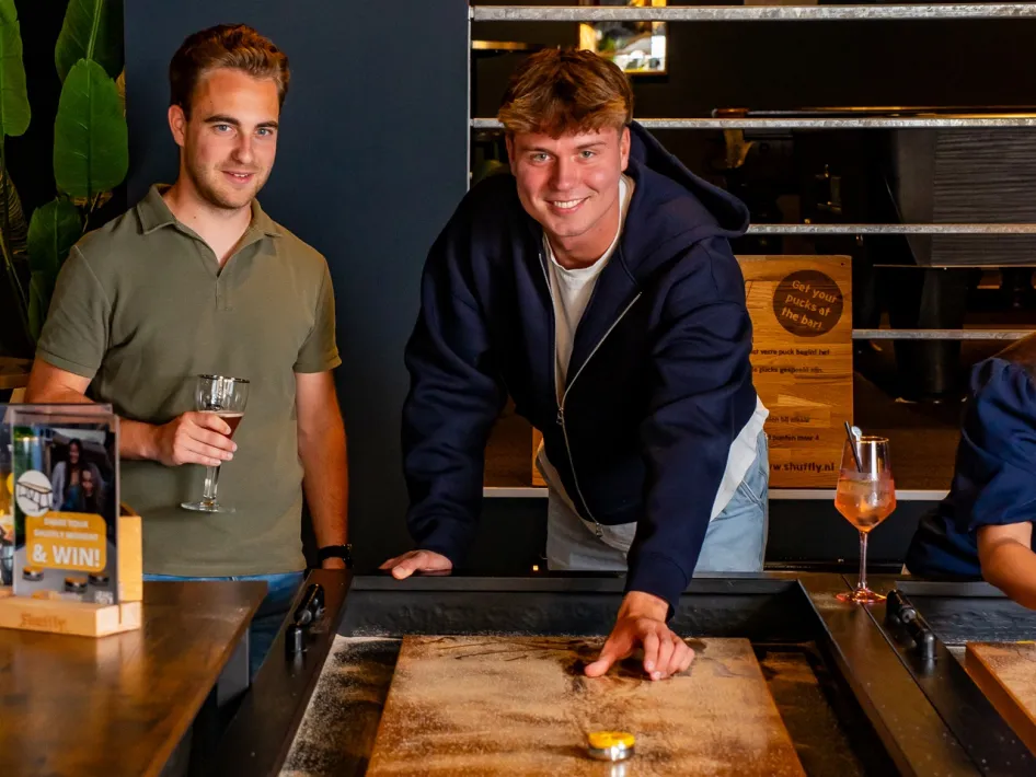 Two men play shuffleboard at a long wooden table while drinks stand along the edge.
