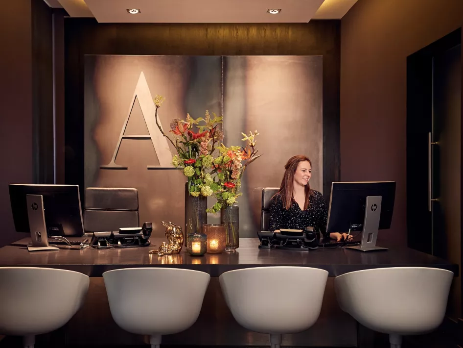 The reception of Het Arresthuis in Roermond, with a welcoming receptionist behind a modern desk, atmospheric lighting and a decorative flower arrangement.