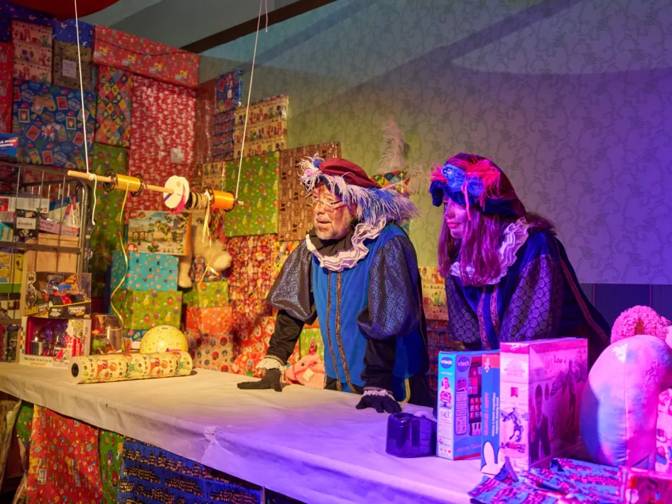 Twee vrolijke Pieten tijdens de Pietenmiddag bijSTOX Roermond, omringd door stapels kleurrijke cadeaus en speelgoed.