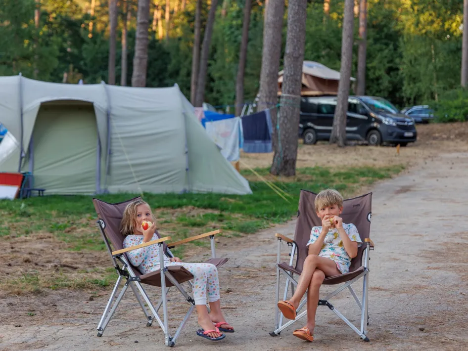 Twee kinderen in pyjama eten fruit op campingstoelen voor een tent tussen de bomen