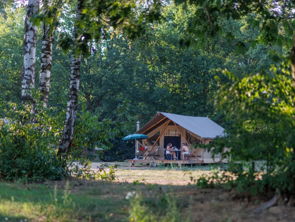 Familie frühstückt an einem Tisch vor einem Glampingzelt von Campingplatz Huttopia de Meinweg, umgeben von Grün und Morgensonne
