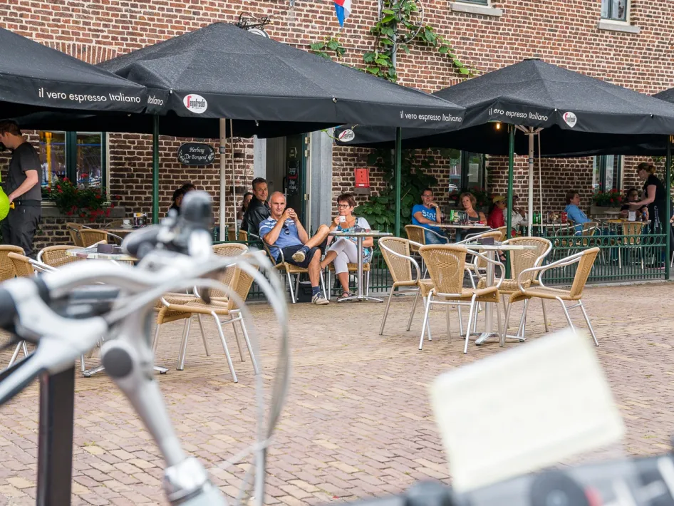 Menschen sitzen unter schwarzen Sonnenschirmen auf der Terrasse am Marktplatz in Beesel, mit Fahrrädern im Vordergrund.