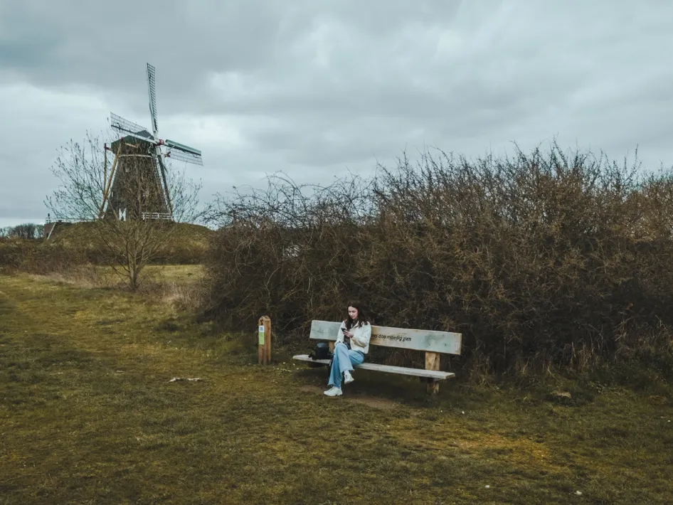 Een vrouw zit op een bankje langs het Huurvaarderspad bij Beesel, met een traditionele windmolen op de achtergrond.