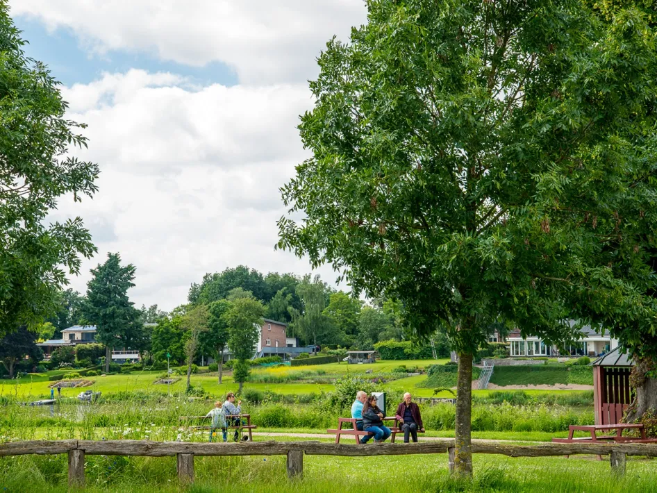 Wanderer ruhen sich vom Huurvaarderspfad auf Bänken in einer grünen Wiese an der Maas in Beesel aus, mit Bäumen und Häusern im Hintergrund.