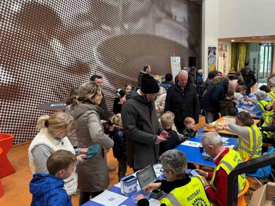Drukke hal waar vrijwilligers van Rotary samen met kinderen en ouders activiteiten begeleiden aan tafels tijdens een evenement.