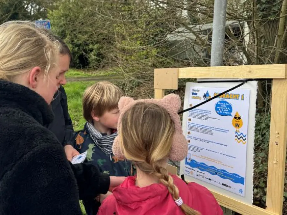 Kinderen en een volwassene bekijken samen een informatiebord met uitleg bij een speurtocht langs een landelijke weg.