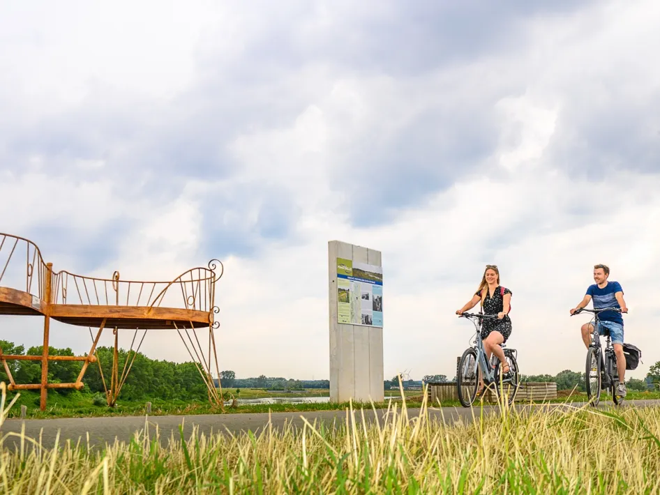 Two cyclists pass an artwork along the International Meuse Cycle Route.