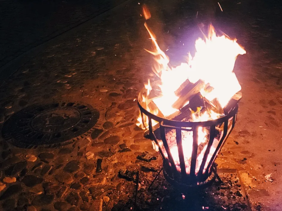 Een knetterend vuur in een metalen vuurkorf op een geplaveid plein, met warme gloed in het donker.