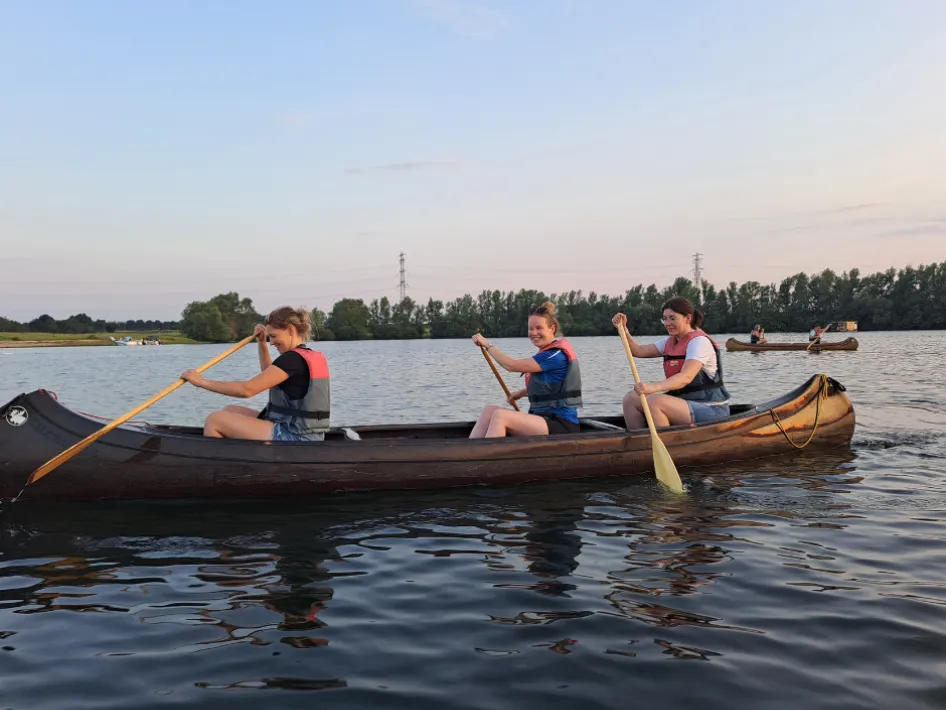 Watersjportiefestasie: Kanotocht -Drie vrouwen kanoën samen op een rustige rivier tijdens zonsondergang, met reddingsvesten aan en peddels in de hand. Op de achtergrond varen nog meer kano’s en is een groene boomrand zichtbaar