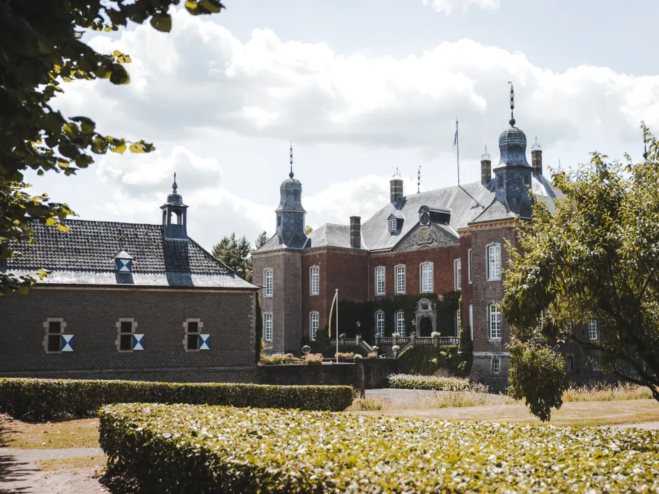 Vooraanzicht op Kasteel Hillenraad met torentjes, schoorstenen en een rijk versierde gevel. Links een bijgebouw met blauwe luiken, omringd door groen en een strak gesnoeide haag.