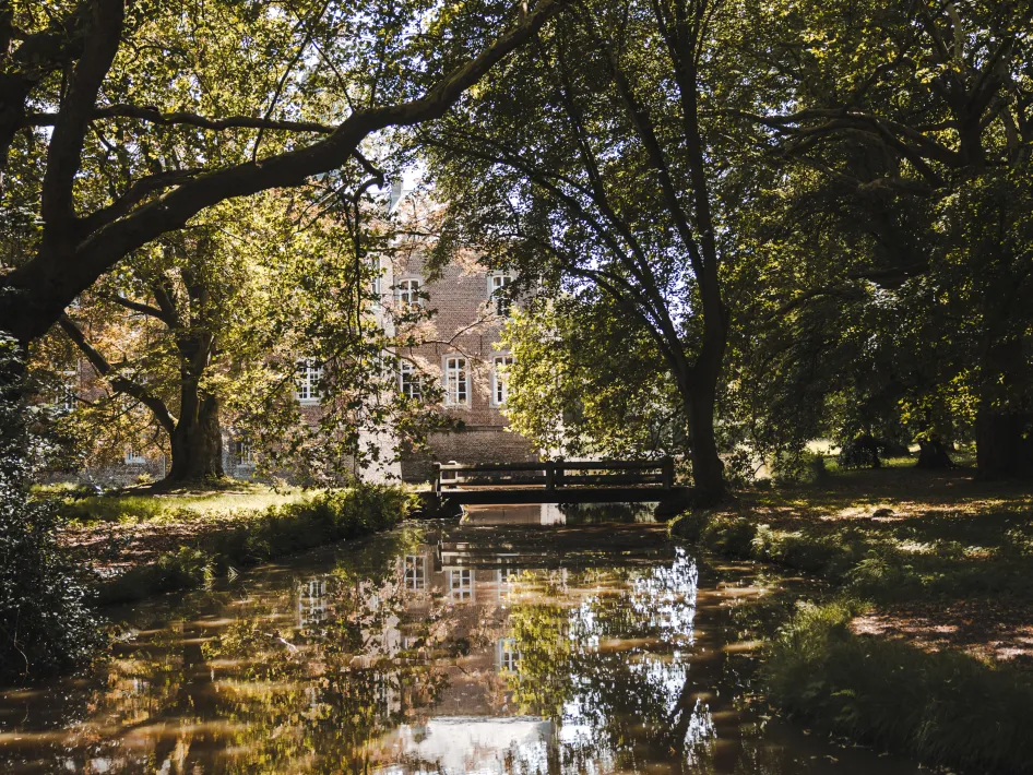 Een stille gracht weerspiegelt de bomen en een bakstenen gevel van Kasteel Hillenraad. Een houten brug verbindt de kasteeltuin met het gebouw, omringd door schaduwrijke bomen.