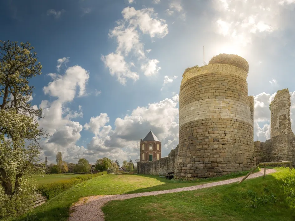 Ruïne van Kasteel Montfort met ronde stenen toren, oude kasteelmuren, gras en wandelpad onder een zonnige wolkenlucht.