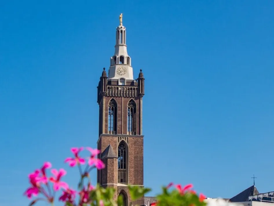 De toren van de Sint-Christoffelkathedraal (Kathedraaltoren) steekt hoog boven Roermond uit tegen een stralend blauwe lucht, met kleurrijke bloemen op de voorgrond.
