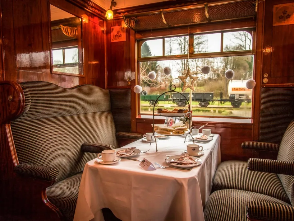 Een feestelijk gedekte tafel met servies en etagère staat in een houten coupé van de Kerst Expres met zicht op het spoor buiten.