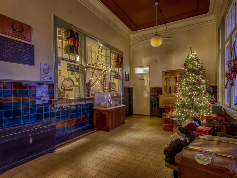 The station hall of Simpelveld with a blue-tiled counter, an illuminated Christmas tree with presents, and two seated dolls on a wooden bench.
