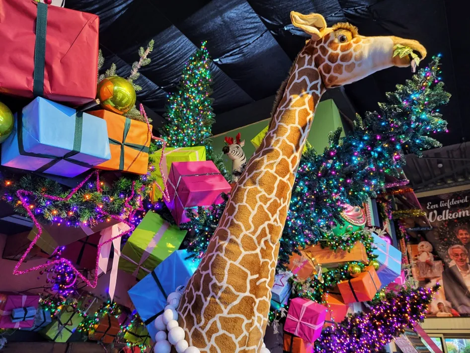 Vrolijke kerstsfeer met een grote pluchen giraf, kleurrijke cadeaus en versierde kerstbomen bij Tuincentrum Daniëls.