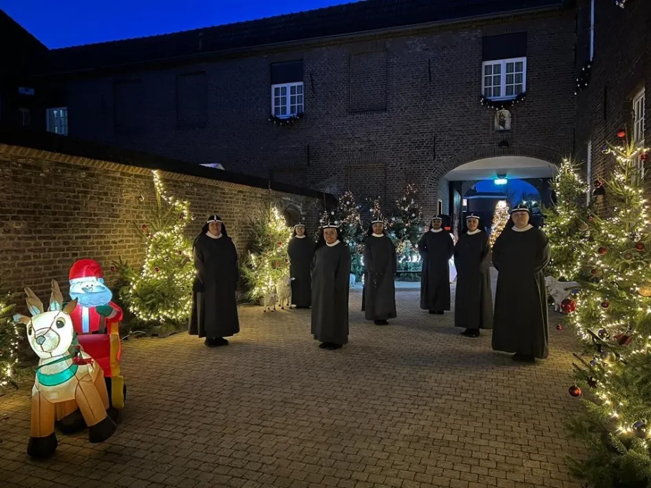 Zusters lopen langs verlichte kerstbomen in de abdijtuin tijdens de Kerststal bij de Zusters in Weert, omgeven door warme lichtjes en kerstdecoraties.