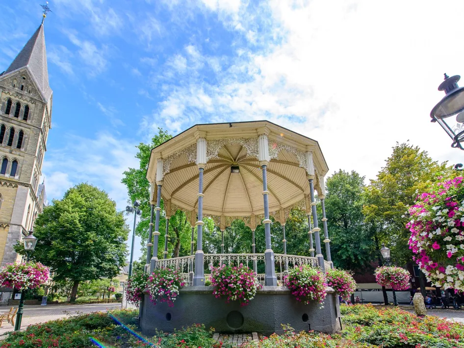 De kiosk op het Munsterplein in Roermond, met gietijzeren zuilen, sierlijk smeedwerk en bloeiende bloemen rondom, naast de toren van de Munsterkerk op een zonnige dag.