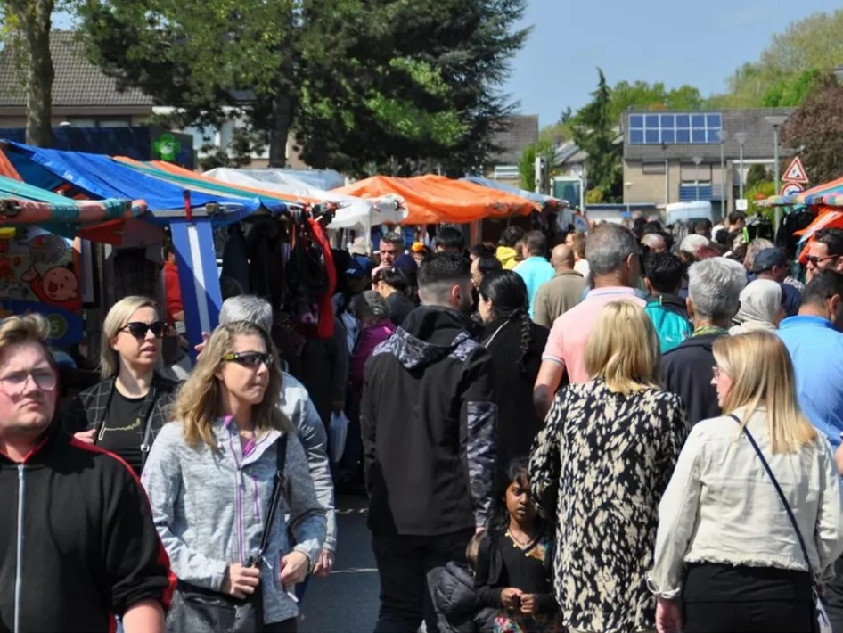 Drukke en gezellige jaarmarkt met kleurrijke kraampjes en een divers publiek dat langs de stands struint op zoek naar koopjes en bijzondere producten.