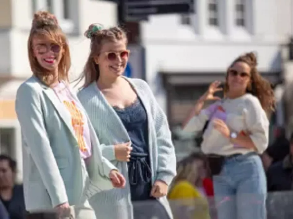 Twee jonge vrouwen poseren lachend in zomerse kleding tijdens een evenement in de stad, met een derde vrouw op de achtergrond.