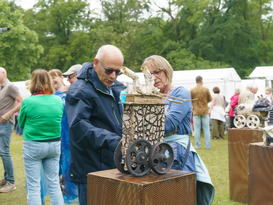 Bezoekers bekijken samen een decoratief metalen object op een sokkel tijdens een markt in de buitenlucht, met andere kramen en mensen op de achtergrond.