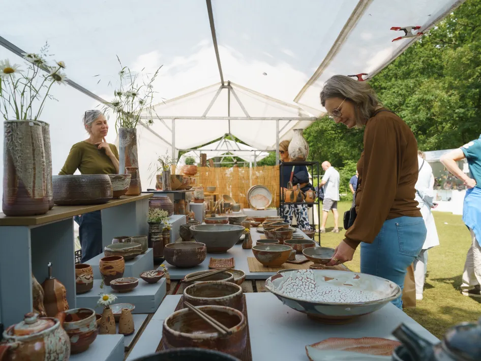 Bezoekers bekijken en kopen handgemaakt keramiek bij kramen onder een witte tent tijdens een markt in de buitenlucht.