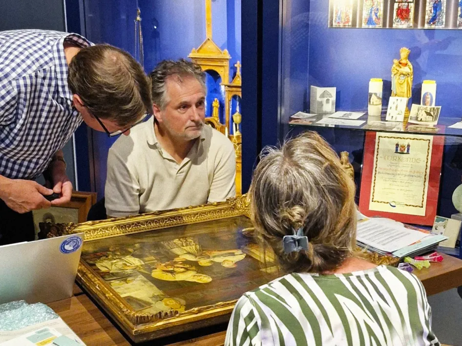 Een man en vrouw laten een religieus schilderij taxeren door een expert in een museumruimte met blauwe vitrines vol erfgoedobjecten.