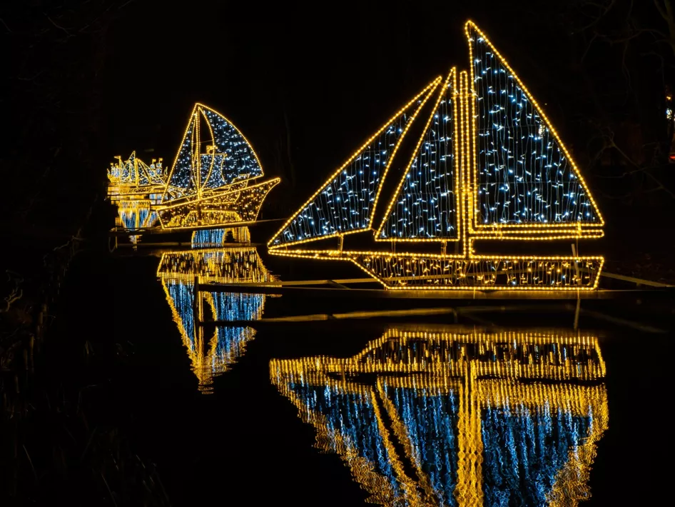 Verlichte bootvormige lampionnen weerspiegelen in het water tijdens de Lampionnentocht in Roermond, sfeervol verlicht in blauw en goud tegen de donkere avondlucht.