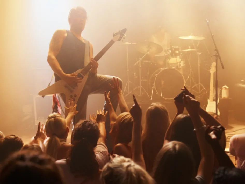 Gitarist op een rockpodium tijdens LEGENDS of ROCK in Wessem, met enthousiaste toeschouwers op de voorgrond en een drummer op de achtergrond in mistig podiumlicht.