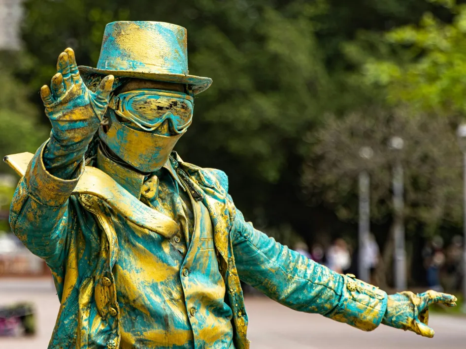 Levend standbeeld in metallic blauw en goud, volledig verkleed met hoed en bril, poseert op een plein met bomen op de achtergrond. Zo'n standbeelden kun je ook zien tijdens het levend standbeeldenfestival in Weert