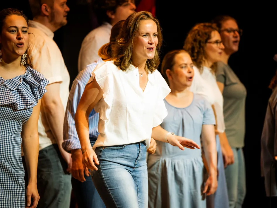 Groep zangers treedt samen op tijdens de Limburgse Koordag, met een zangeres in een witte blouse centraal op het podium.