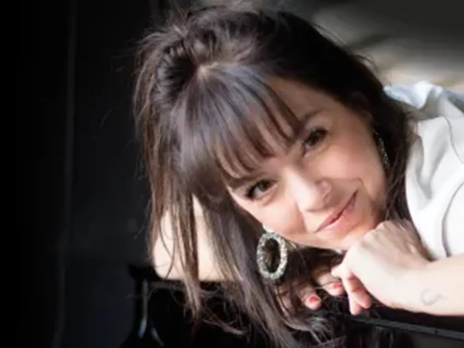 Portret van vrouw met lange donkere haren en oorbellen die glimlachend leunt, promotiebeeld voor een concert