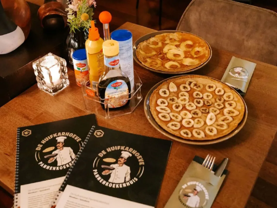 Pfannkuchen mit Obst auf einem Tisch im Pannenkoekenhuis de Huifkarhoeve, mit Speisekarten und Geschirr.