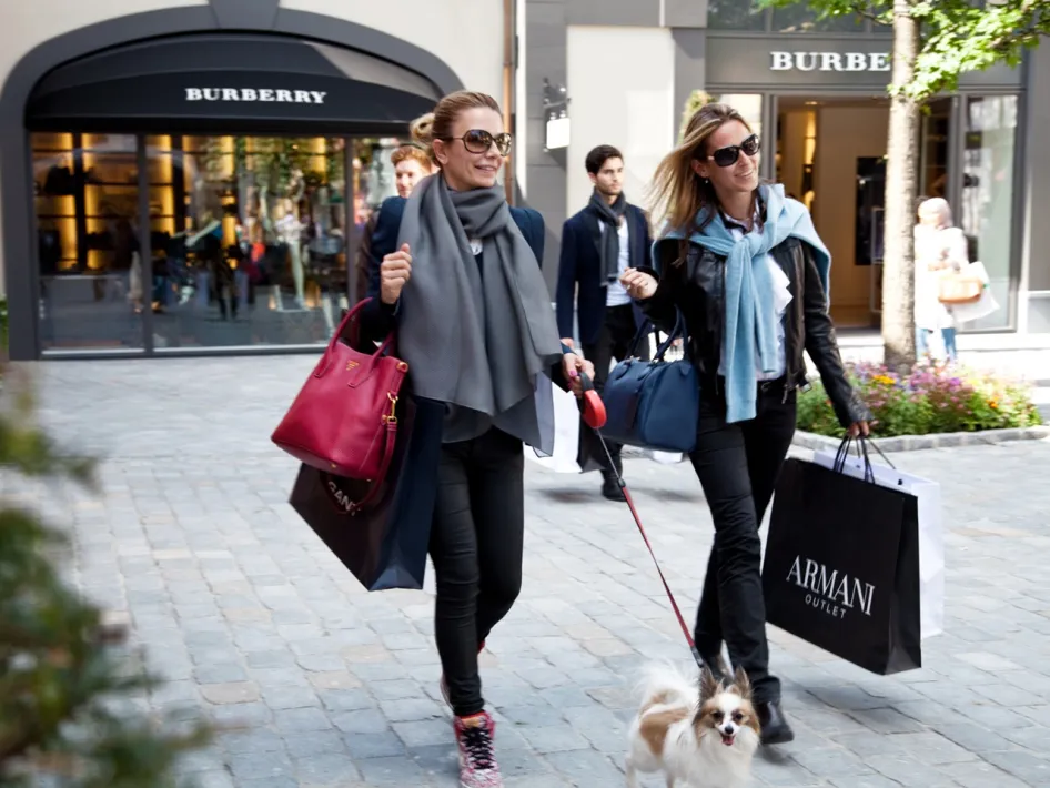 Twee vrouwen met luxe shoppingtassen en een hondje wandelen tijdens de Luxury Days bij Designer Outlet Roermond langs de winkels van Burberry en Armani.