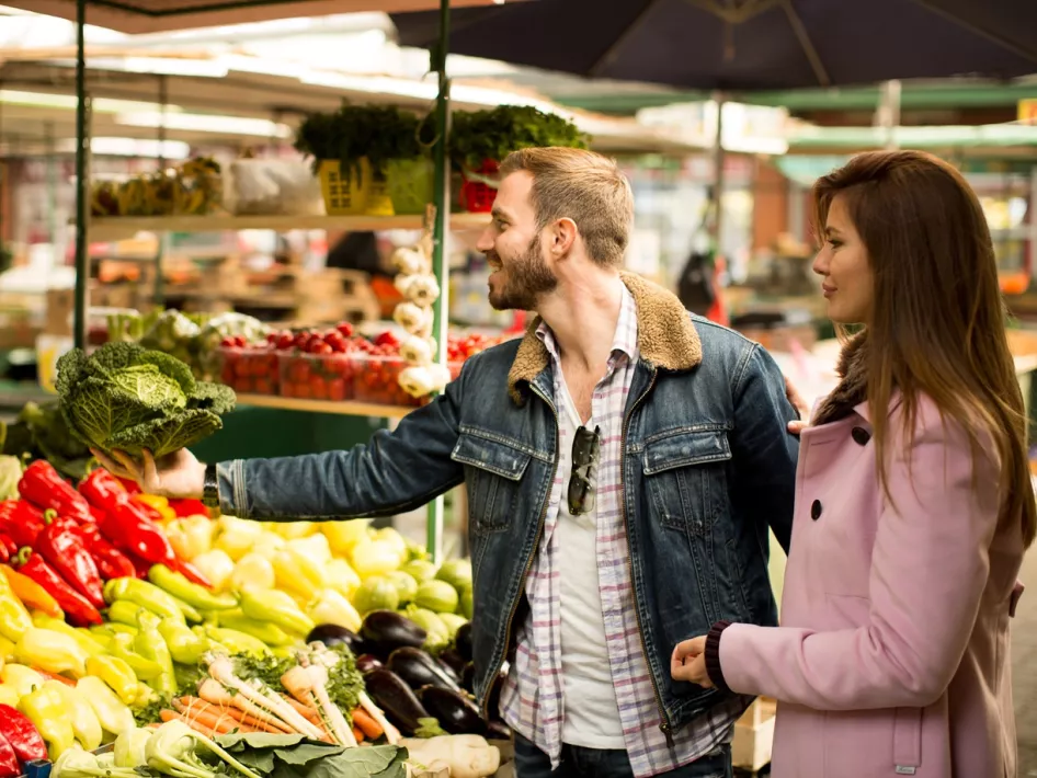 Man en vrouw bekijken verse groenten op een marktstand met paprika’s, kool, wortels en aubergines.