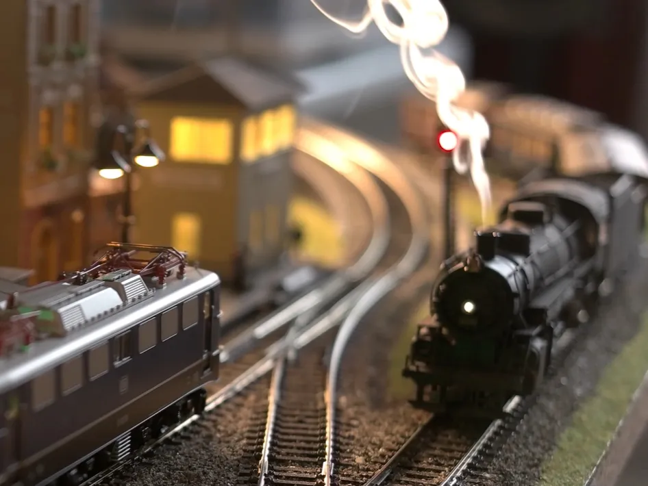 Modeltreinen op een modelspoorbaan tijdens een modeltreinenbeurs, met rook, verlichting en miniatuurhuisjes.