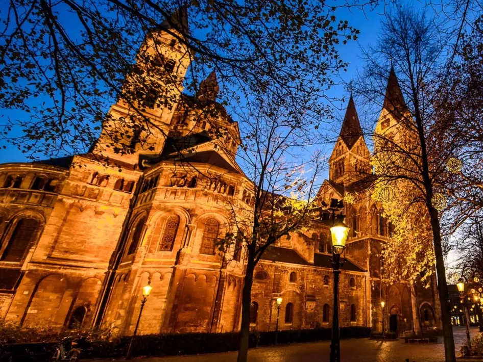 Verlichte kathedraal in avondlicht, omringd door bomen en straatlantaarns. De imposante architectuur en sfeervolle verlichting geven het plein een warme, historische uitstraling.