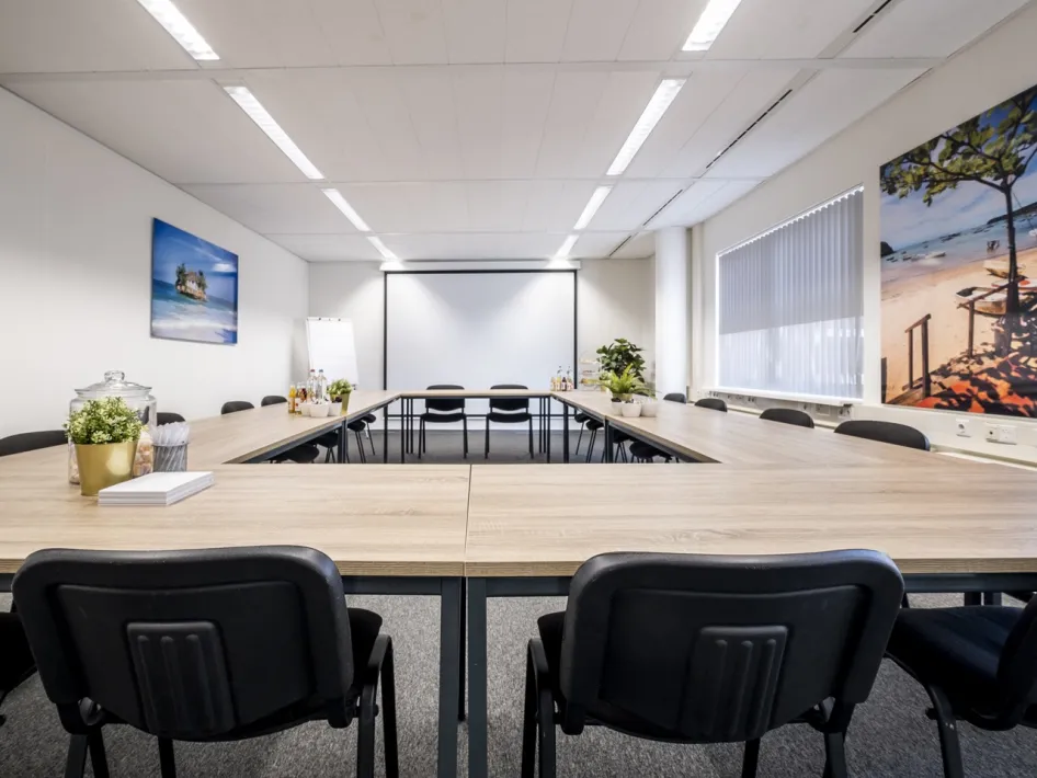Vergaderruimte in U-opstelling met presentatiescherm en daglicht, ingericht voor zakelijke bijeenkomsten.