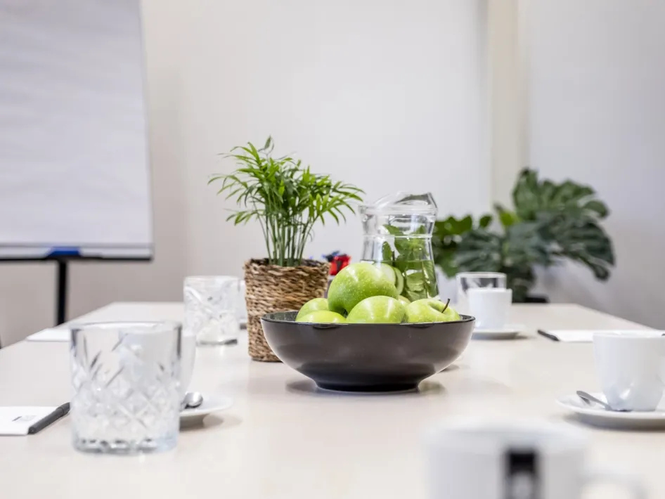 Detailaufnahme eines Besprechungstisches mit Gläsern, Äpfeln, Pflanzen und Notizblöcken in einem hellen Raum.