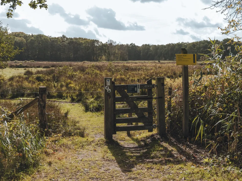 Houten toegangshek met informatiebord bij het Wijffelterbroek, een van de natuurgebieden van het KempenBroek, met grasland en bomen op de achtergrond.