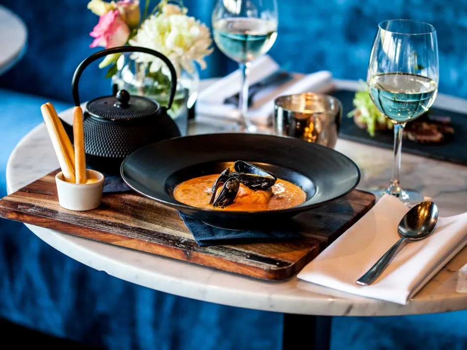 Ein tiefschwarzes Teller mit einer cremigen Muschelsuppe, serviert auf einem Holzbrett mit Grissini, Weißwein und einer schwarzen Teekanne auf einem stimmungsvoll gedeckten Tisch.