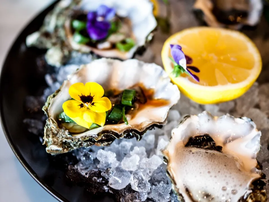 A plate of fresh oysters on a bed of ice, garnished with edible flowers, spring onions and half a lemon.
