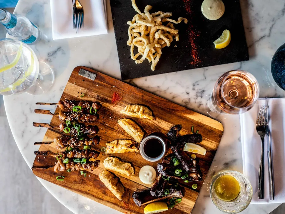Ein Holzbrett voller verschiedener Spieße, frittierter Häppchen und Dip-Saucen, serviert mit Rosé und Sprudelwasser auf einem Marmortisch.