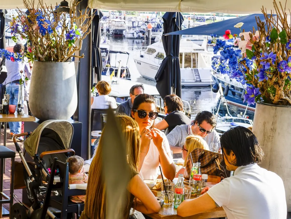 A lively atmosphere on the terrace at Grand Café NUby the water, with people seated at tables, flowers in large pots and a view of moored boats.