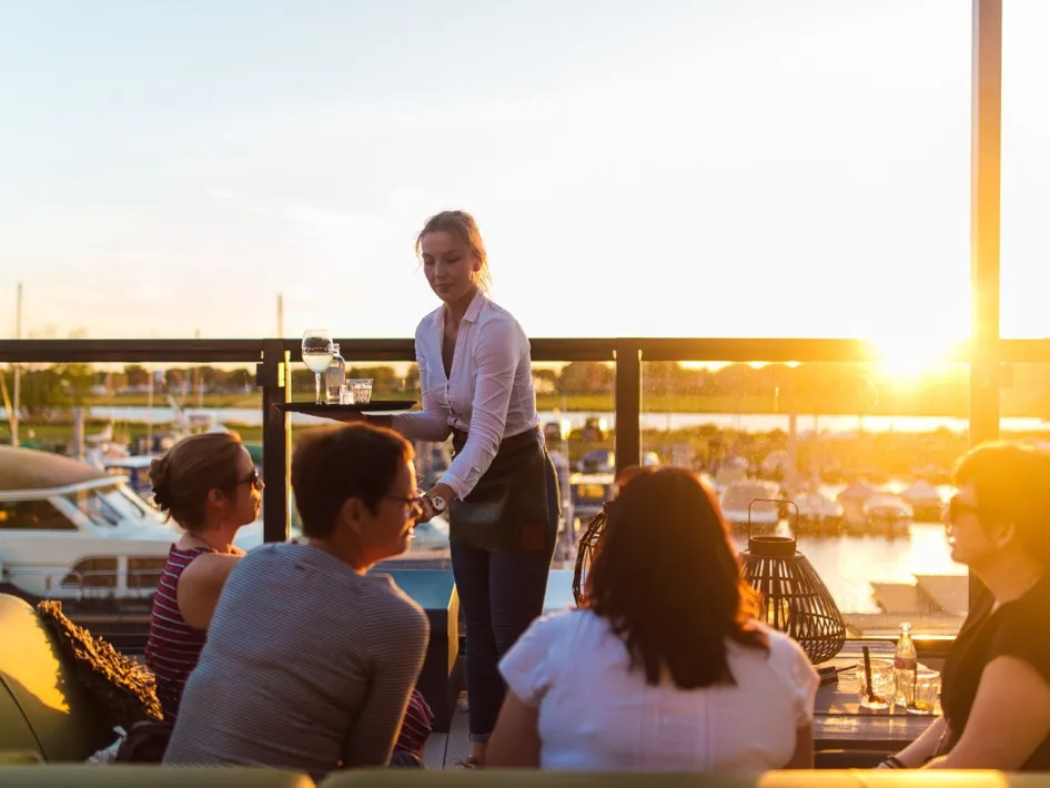 Eine Kellnerin bringt einer Gruppe von Gästen auf einer Dachterrasse Getränke, mit Blick auf den Yachthafen bei Sonnenuntergang.