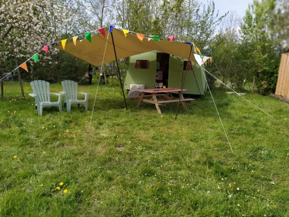 Gemoedelijke kampeerplek met retrocaravan, kleurrijke vlaggetjes en picknicktafel op een grasveld, met bloeiende bomen op de achtergrond.