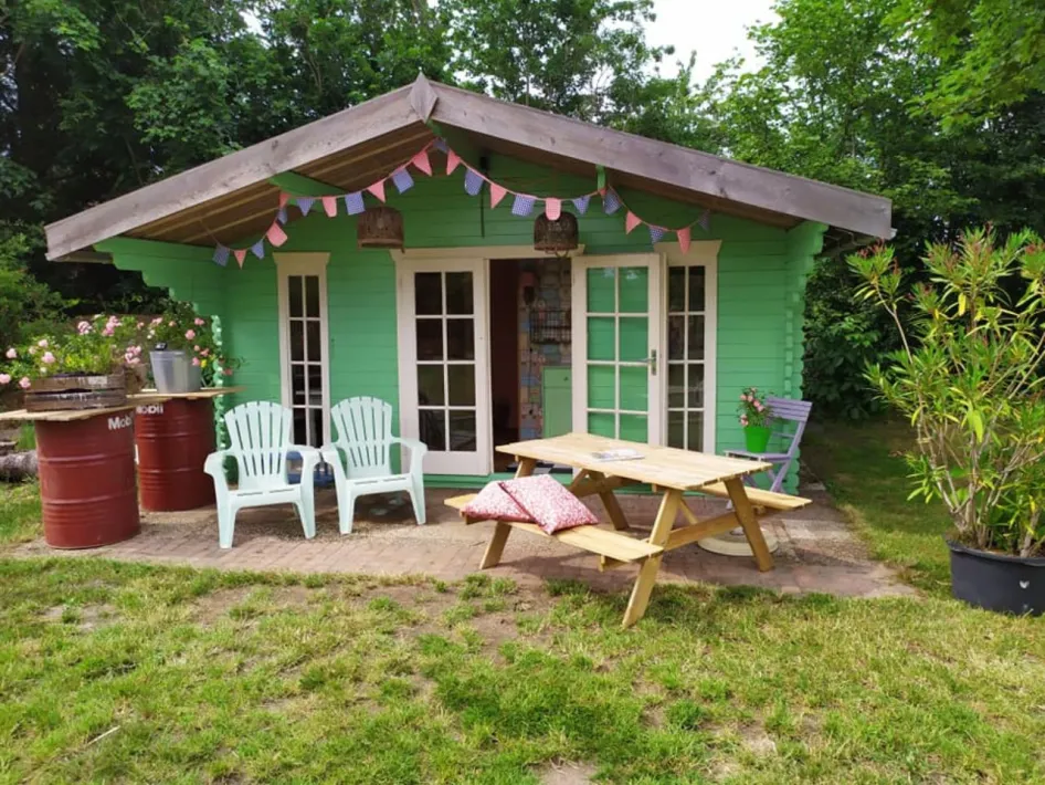 Lichtgroen houten vakantiehuisje met picknicktafel en stoelen, vrolijk versierd met slingers en omgeven door bomen en planten.
