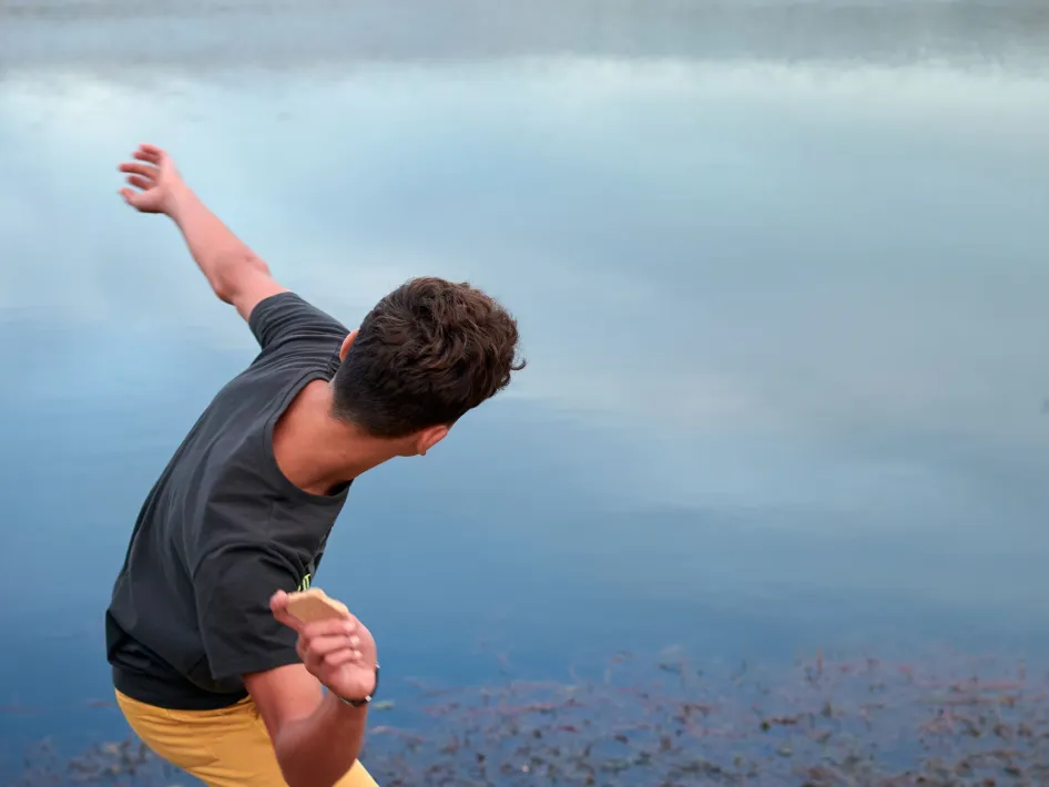 Persoon die een platte steen over het water probeert te ketsen tijdens Stoneskimming, met zicht op een rustige waterplas.