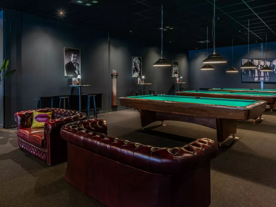 Ein stilvoller Billardsaal mit mehreren Pooltischen, stimmungsvoller Beleuchtung und luxuriösen Ledersofas. An den Wänden hängen Schwarz-Weiß-Porträts, die zur klassischen Atmosphäre beitragen.